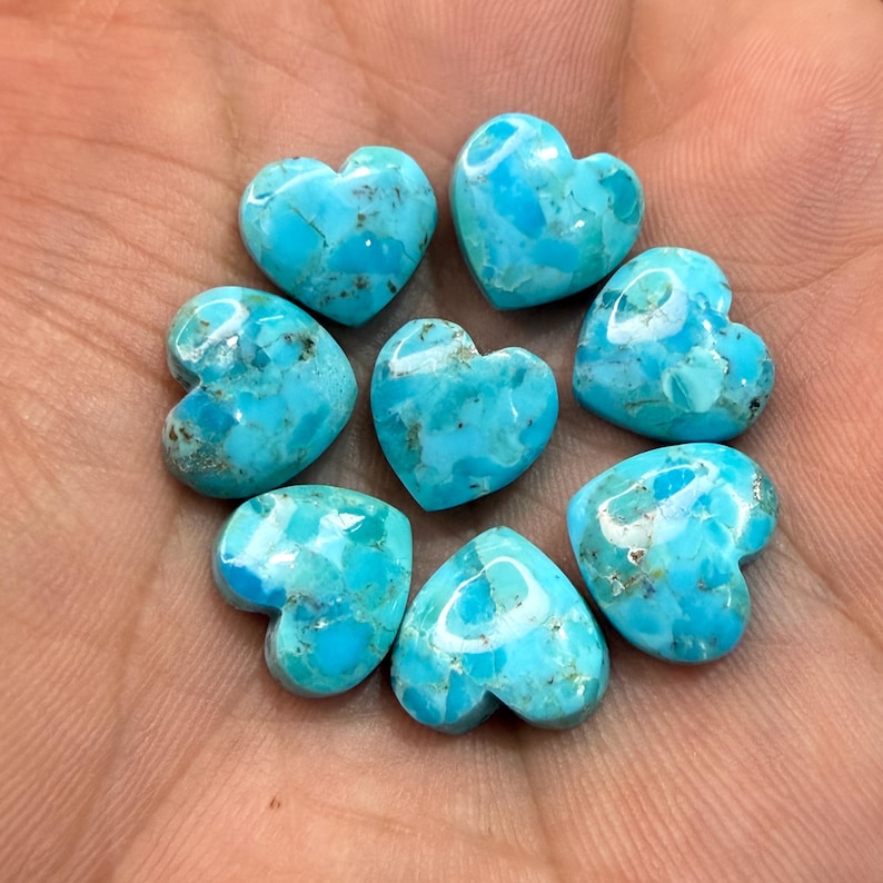 May include: Seven polished heart-shaped turquoise gemstones arranged in a circular pattern. Each stone features a vibrant sky-blue color with natural dark brown matrix veining throughout the smooth, glossy surfaces of the heart-shaped cabochons.