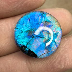 May include: A round, iridescent gemstone with a vibrant display of blues, greens, and purples. The stone features a cracked, web-like pattern across its surface, with areas of deep black and a bright, reflective sheen.