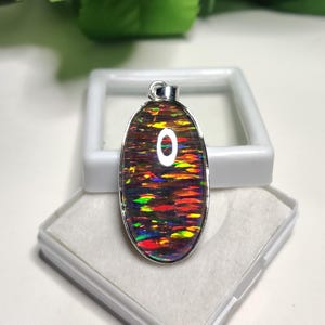 Może przedstawiać: Srebrny wisiorek z kolorowym opalem, który prezentuje tęczę kolorów, w tym czerwony, pomarańczowy, żółty, zielony, niebieski i fioletowy. Opal jest osadzony w srebrnej owalnej oprawie.