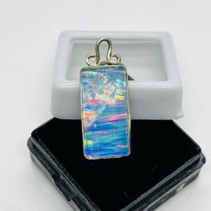 May include: A silver pendant with a rectangular opal stone. The opal is iridescent with shades of blue, pink, and white.