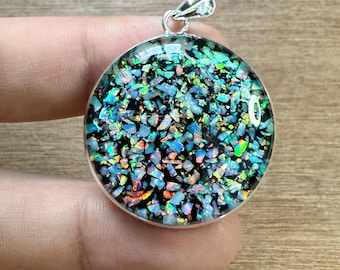 May include: A round pendant with a silver bail. The pendant features a mosaic of iridescent, colorful pieces set against a black background. The colors include blues, greens, and hints of orange.