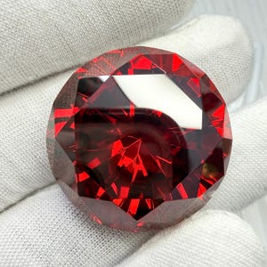 May include: A large, faceted, round gemstone with a deep red color. The gem has many reflective surfaces, creating a brilliant display of light. The gem is held by a white gloved hand.