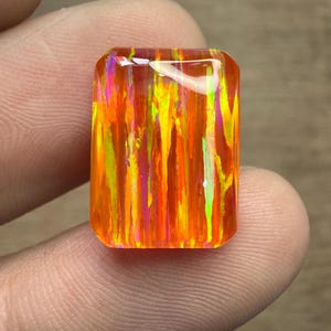 May include: A rectangular gemstone with a vibrant, fiery color palette. The stone features vertical streaks of orange, yellow, pink, and green, creating a dynamic and eye-catching appearance. The gemstone is held between fingers, showcasing its polished surface.