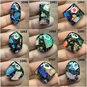 May include: A collection of nine polished gemstones with a mosaic-like appearance. Each stone features a dark base color with embedded fragments of blue, green, orange, and white, creating a sparkling effect. The stones are in various shapes, including oval, rectangular, and square.