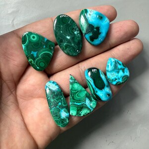 May include: A collection of polished gemstones in various shapes, including oval, teardrop, and irregular forms. The stones display a mix of deep green and turquoise blue colors with intricate patterns. The gemstones are held in a hand.