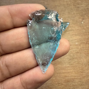 Könnte beinhalten: Eine Hand hält eine durchscheinende, blau-grüne Pfeilspitze. Die Pfeilspitze hat eine scharfe Spitze und gezackte Kanten, mit einem etwas dunkleren Farbton an der Oberseite. Das Licht fängt das glasartige Material ein und erzeugt einen schimmernden Effekt.