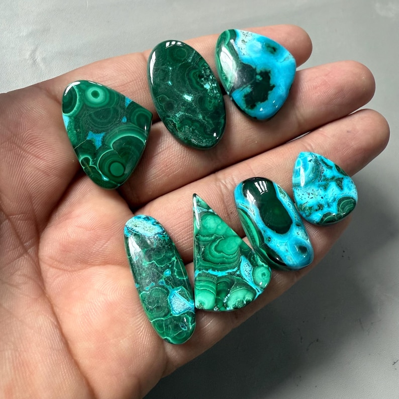 May include: A collection of polished gemstones in various shapes, including oval, teardrop, and irregular forms. The stones display a mix of deep green and turquoise blue colors with intricate patterns and textures. The gemstones are held in a hand.