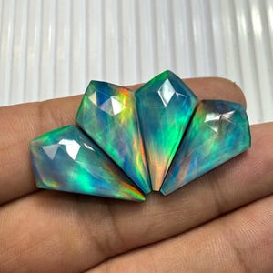 May include: Three kite-shaped faceted gemstones displaying a vibrant play-of-color with iridescent flashes of neon green, electric blue, and warm orange hues. The stones feature a polished, geometric cut with sharp angles and a reflective surface.