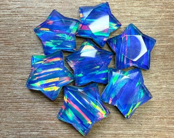 May include: A collection of eight iridescent blue star-shaped gemstones. The faceted stars have a range of colors, including blue, green, and pink, and are arranged on a light brown wooden surface. The stars are a decorative element.