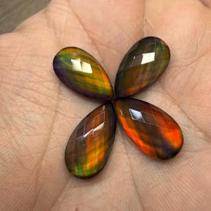 May include: Four faceted, teardrop-shaped gemstones arranged in a flower-like pattern. The gems display a vibrant array of colors, including green, orange, and purple, with a reflective, polished surface. The gems are held in a hand.