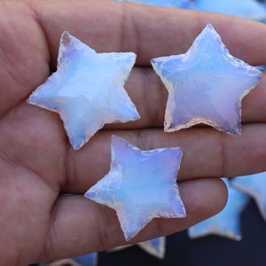 以下が含まれることがあります： 手のひらに置かれた、虹色の青い星型のオパール石が3つ。石は半透明で、乳白色をしています。