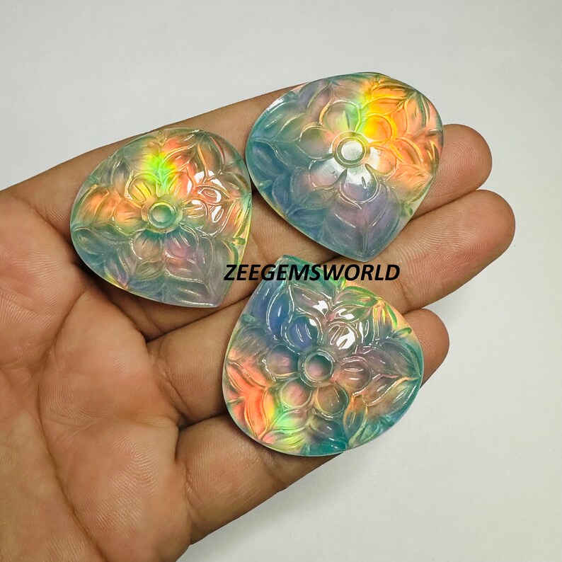 May include: Three iridescent gemstone cabochons carved with a floral design. The cabochons are a light blue color with rainbow iridescence.