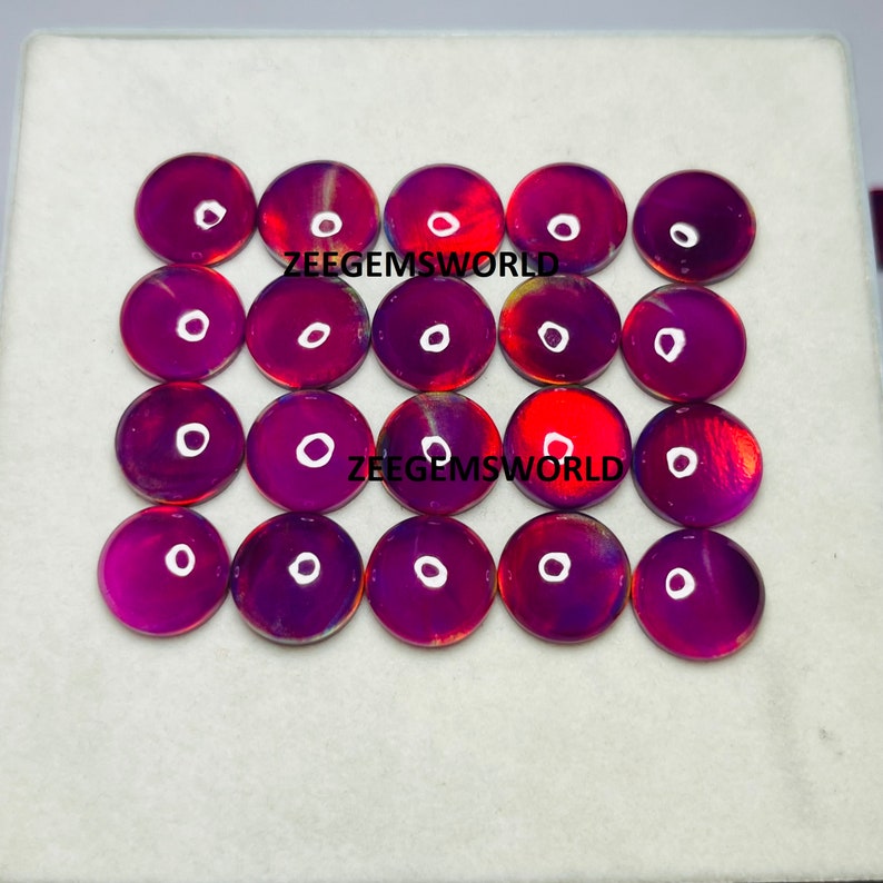 May include: Twenty round, iridescent cabochons in shades of purple, red, and gold. The cabochons are arranged in a grid pattern on a white surface. The text "ZEEGEMSWORLD" appears twice on the surface.