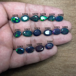 Puede incluir: Una colección de gemas ovaladas en varios tonos de verde, azul y negro, numeradas del 1075 al 1090. Las gemas se muestran en una mano, mostrando sus diferentes colores y patrones. Es probable que sean ópalos.