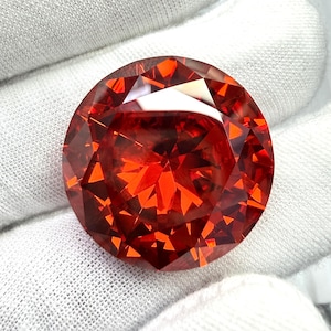 May include: A large, round, faceted gemstone with a vibrant red-orange hue. The gem's intricate cuts create a dazzling display of light and brilliance. The gemstone is set against a white background.
