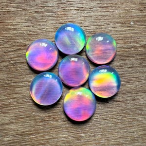 Puede incluir: Siete piedras preciosas redondas e iridiscentes con un acabado pulido. Las piedras muestran una gama de colores, incluyendo rosa, morado, azul y verde, con una calidad brillante y etérea. Las piedras están dispuestas en un patrón circular.