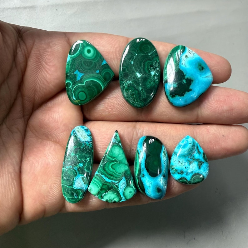 May include: A collection of polished gemstones in various shapes, including oval, teardrop, and triangular. The stones display a mix of vibrant green and turquoise colors with unique swirling patterns, ideal for jewelry making or collecting.