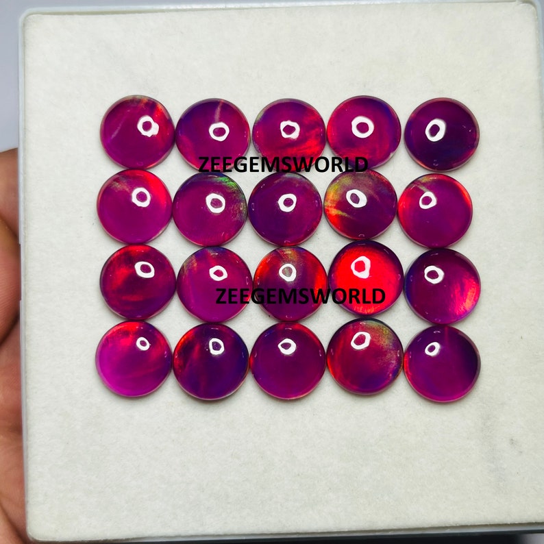 May include: Twenty round, iridescent opal cabochons in shades of purple, red, and gold. The cabochons are arranged in four rows of five, with the text "ZEEGEMSWORLD" printed twice on the white surface below.
