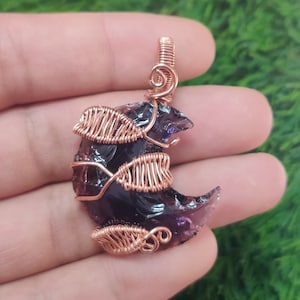 Op de afbeelding: Een halve maanvormige hanger gemaakt van een stuk amethist met koperdraad eromheen gewikkeld. De draad is gevormd in bladeren en spiralen.