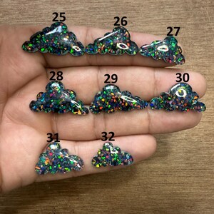May include: A collection of cloud-shaped resin cabochons with iridescent glitter. Each cloud is a different size and shape, with numbers 25 through 32 printed on them. The glitter reflects a variety of colors, including green, blue, and orange.