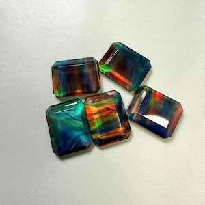 Puede incluir: Cinco piedras preciosas rectangulares con bordes biselados. Las piedras muestran una vibrante gama de colores, incluyendo azules, verdes, naranjas y rojos, creando un efecto arcoíris. Las gemas están dispuestas sobre una superficie blanca.
