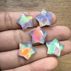 Può includere: Sei gemme iridescenti a forma di stella. Ogni stella ha un design sfaccettato, che mostra uno spettro di colori tra cui rosa, verde e arancione. Le gemme sono poste su uno sfondo neutro, evidenziando il loro aspetto vibrante e multicolore.