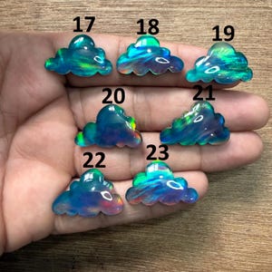 May include: A hand holding seven cloud-shaped gemstones. The gemstones are numbered 17 through 23. Each gemstone has a unique color pattern, with hues of blue, green, and hints of other colors. The gemstones are translucent and appear to be polished.