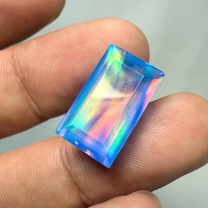 Puede incluir: Una gema rectangular e iridiscente con un tono azul y un corte rectangular. La gema muestra un espectro de colores, incluyendo rosa, amarillo y verde, que reflejan la luz. La piedra se sostiene entre los dedos.
