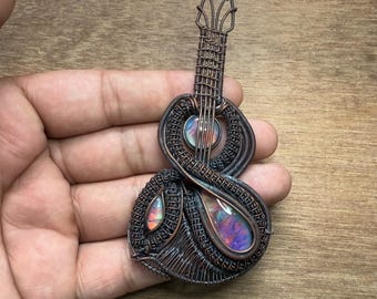 Puede incluir: Colgante de alambre de cobre hecho a mano con forma de mandolina. El colgante presenta tres piedras de ópalo iridiscentes en tonos morado, naranja y azul. El alambre está intrincadamente tejido para crear el cuerpo y el mástil del instrumento.