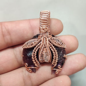 Op de afbeelding: Een hanger van koperdraad met een donkerpaarse edelsteen. De hanger heeft een delicaat, ingewikkeld ontwerp met een lus aan de bovenkant om op te hangen.