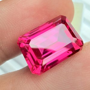 May include: A faceted, rectangular-cut, vibrant pink gemstone. The gem has a high degree of clarity, with multiple facets reflecting light. The color is a deep, saturated pink, with a slight variation in tone across the surface.
