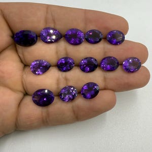May include: A collection of polished, faceted amethyst gemstones in various shapes, including oval and pear cuts. The gemstones are a deep, rich purple color and are displayed against a neutral background.