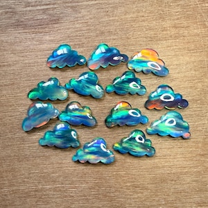 May include: A collection of iridescent cloud-shaped gemstones in various shades of blue, green, and orange. The gemstones are arranged on a wooden surface, showcasing their unique color variations and reflective qualities. These could be used for jewelry making or other crafts.
