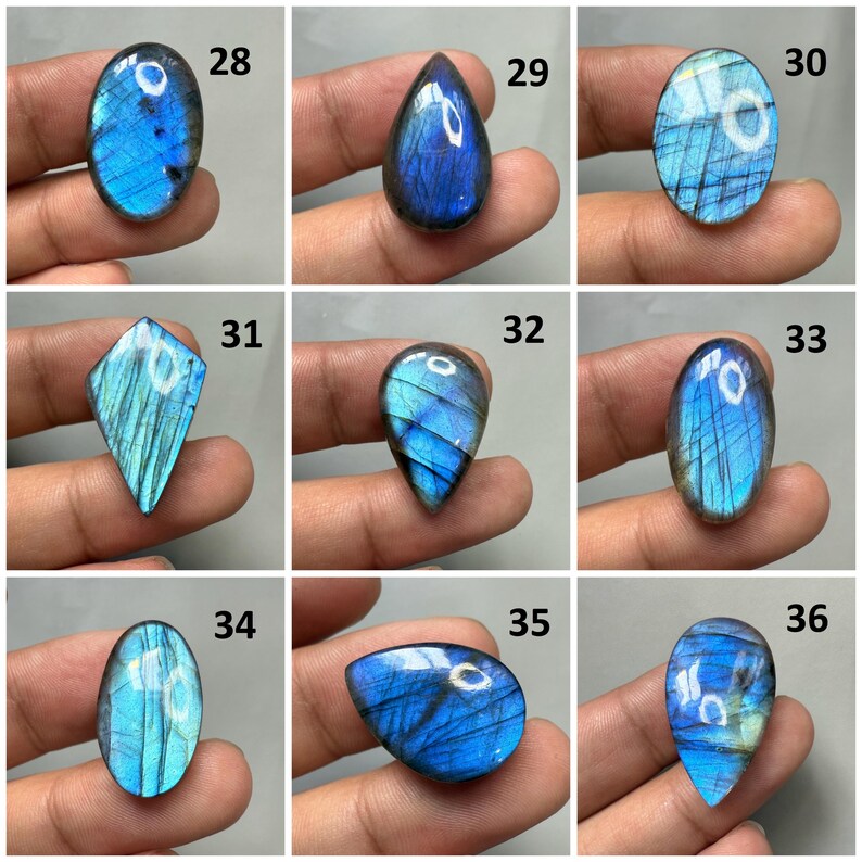 Op de afbeelding: Negen gepolijste labradoriet edelstenen in verschillende vormen, waaronder ovaal, druppel en ruit. De stenen vertonen iriserende blauwe en teal flitsen. Elke steen wordt in een hand gehouden, met de nummers 28 tot en met 36 zichtbaar.