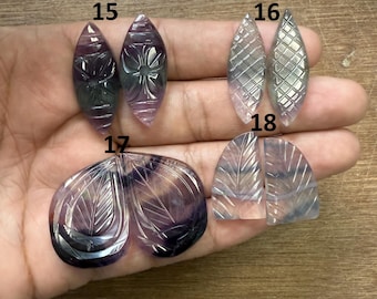 Puede incluir: Una mano muestra cuatro pares de cabujones de piedras preciosas talladas. Las piedras tienen varias formas, incluyendo diseños de lágrima y hoja. Las piedras son moradas, grises y transparentes con patrones florales y geométricos tallados. Las piedras preciosas están numeradas 15, 16, 17 y 18.