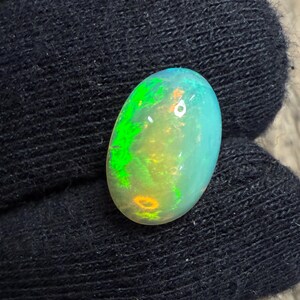 Könnte beinhalten: Ein ovaler Opal-Edelstein mit einem lebendigen Farbspiel. Der Stein hat eine Basis aus hellblauen und gelben Farbtönen mit auffälligen grünen Blitzen. Die polierte Oberfläche reflektiert das Licht und zeigt das innere Farbspiel.