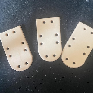 May include: Three light tan leather pieces with rounded bottoms and multiple evenly spaced holes. The pieces are arranged on a dark surface, likely for crafting or leatherwork projects.