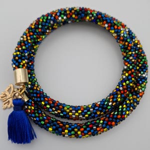 Könnte beinhalten: Ein kreisförmiges Perlenarmband aus einem durchgehenden Strang kleiner, mehrfarbiger Glasperlen in Blau-, Grün-, Gelb-, Orange- und Silbertönen. Ein goldfarbener Metallverschluss verbindet die Enden und ist mit einem kleinen goldenen Schmetterlingsanhänger sowie einer leuchtend blauen Stoffquaste versehen.
