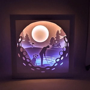 May include: A white paper silhouette of a golfer on a golf course with a golf cart and a moon in the background. The silhouette is lit from behind, creating a glowing effect.