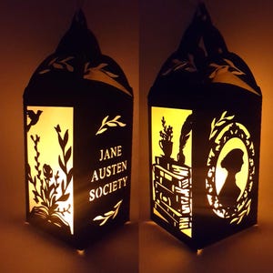 May include: Black paper lantern with cut-out designs and the words "Jane Austen Society." The lantern features silhouettes of flowers, books, a quill, and a portrait. The interior glows with a warm light, creating a cozy ambiance.