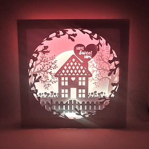 Może przedstawiać: Biała lampka z papieru z czerwonym światłem oświetla wycięty wzór domu z ogrodzeniem, drzewami i napisem "Home Sweet Home" w kształcie serca.