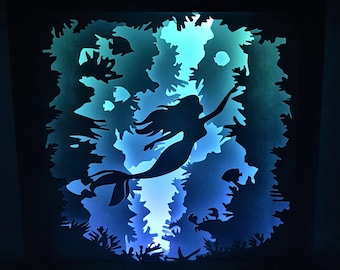 May include: A blue and green light illuminates a paper cut-out of a mermaid swimming through a sea of coral. The mermaid is silhouetted against the light.