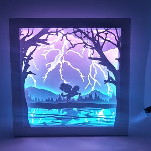 May include: A white frame with a paper cut-out design of a winged creature flying over a lake with lightning strikes in the background. The frame is illuminated with purple and blue lights.