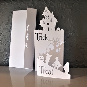 May include: White paper card with a cut-out design of a haunted house, a tree, a ghost, a witch, and a pumpkin. The card says "Trick or Treat" in black lettering.