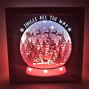 May include: A white wooden frame containing a lit-up paper cut-out design of a snow globe with Santa Claus and reindeer flying over a snowy forest. The text "Jingle All The Way" is written around the top of the snow globe.