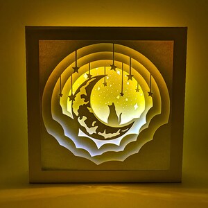 May include: A layered paper art piece featuring a cat silhouette on a crescent moon, surrounded by stars. The artwork is illuminated with a warm yellow glow, creating a dreamy, celestial scene. The piece is framed in a white square.