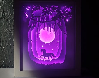 Puede incluir: Una obra de arte de papel recortado enmarcada en blanco con una silueta de unicornio en una escena de bosque. La escena está iluminada con una luz morada, creando un efecto mágico.