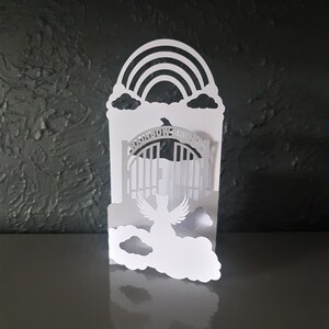 May include: White paper cut-out of a rainbow, clouds, a gate with the words "Rainbow Bridge" and an angel with wings flying through the gate.
