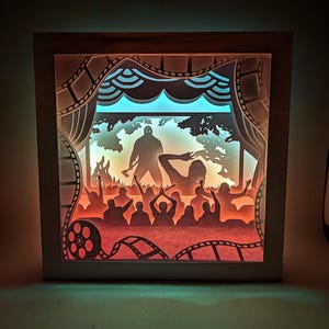 Könnte beinhalten: Ein mehrschichtiges Papierkunstwerk, das eine Horrorfilmszene darstellt. Das Kunstwerk zeigt eine Silhouette mit einer Waffe, eine Menschenmenge und Filmelemente, alles in einem beleuchteten Rahmen. Die Farbpalette umfasst Orange-, Rot- und Blautöne.