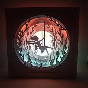 May include: A white papercraft lamp with a fairy silhouette on a swing in a woodland scene. The lamp is illuminated with a multi-coloured light.